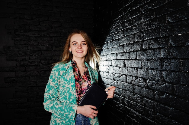 Стильная блондинка в куртке и джинсах с блокнотом в руках у кирпичной черной стены в студии