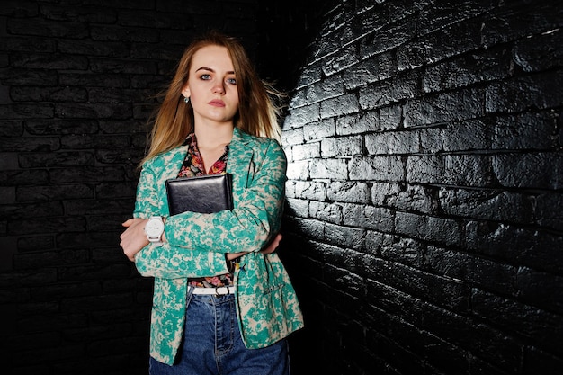 Stylish blonde girl in jacket and jeans with notebook diary at hands against brick black wall at studio