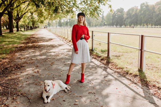 Stylish blonde girl wearing black hat and white skirt posing with her dog on the park path.