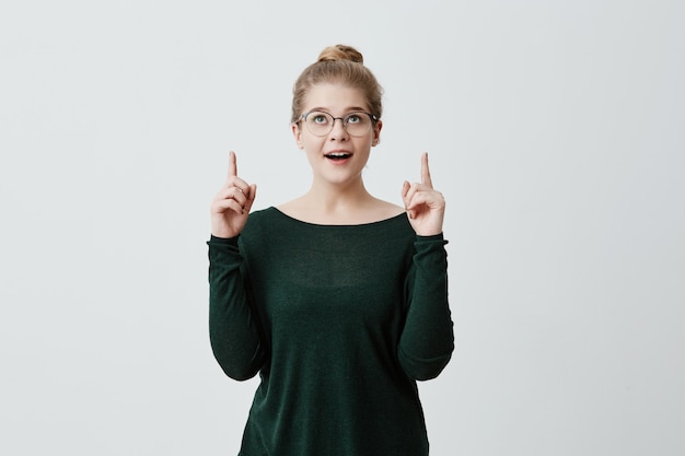 Stylish blonde girl with eyeglasses on, popped eyes, raises fore fingers with excitement, being glad and surprised, has good mood. People emotions and feelings. Face expressions