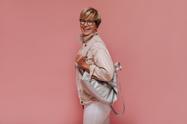 Стильная блондинка с короткими волосами в очках в бежевой прохладной куртке и белых брюках улыбается и держит сумку на розовом фоне.