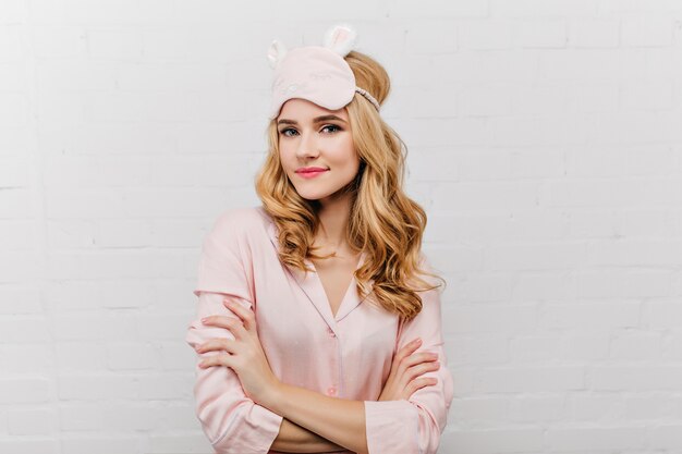 Stylish blue-eyed woman in silk pajama posing with arms crossed. Fascinating curly girl in sleep mask standing in confident pose.