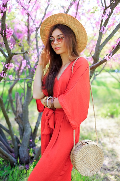 Stylish bohemian girl posing in coral dress and straw hat near blossom cherry tree in spring park.