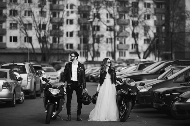 세련된 신부와 수염 난 신랑은 도시 거리의 아름다운 쿠데타에서 스포츠 오토바이 옆에 서 있습니다...