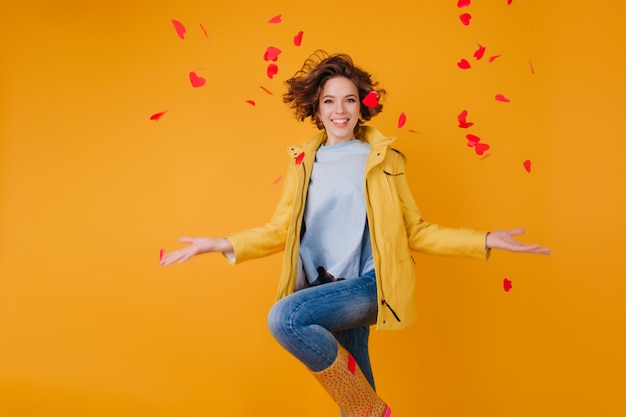 Stylish brown-haired girl throwing out hearts while jumping on yellow wall. Pretty european young woman in jacket having fun in valentine's day.