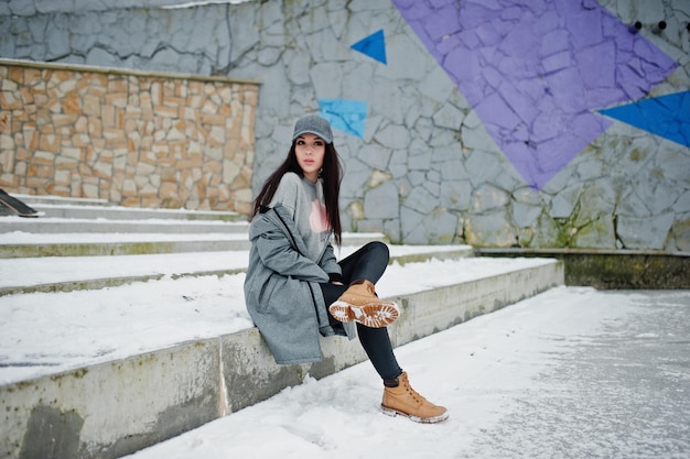 회색 모자 캐주얼 스트리트 스타일의 세련된 브루네트 소녀는 겨울날 색깔의 벽에 기대어