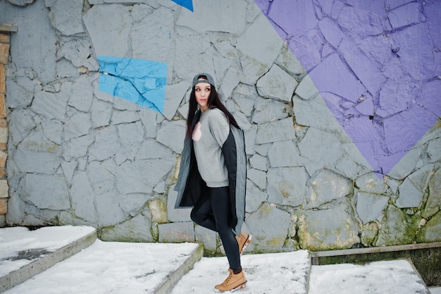 Stylish brunette girl in gray cap casual street style on winter day against colored wall