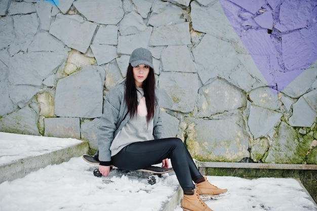 Stylish brunette girl in gray cap casual street style with skate board on winter day against colored wall