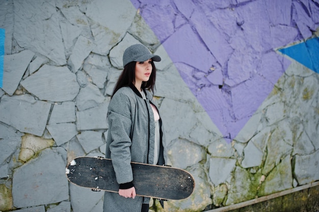 Stylish brunette girl in gray cap casual street style with skate board on winter day against colored wall