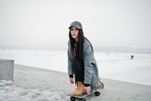 Стильная брюнетка в серой кепке в повседневном уличном стиле со скейтбордом в зимний день