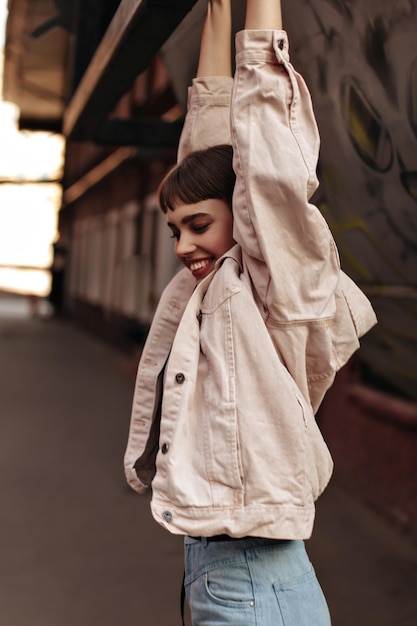 Stylish brunette girl smiling outdoors Positive woman in light denim jacket and cool jeans has fun and poses at street