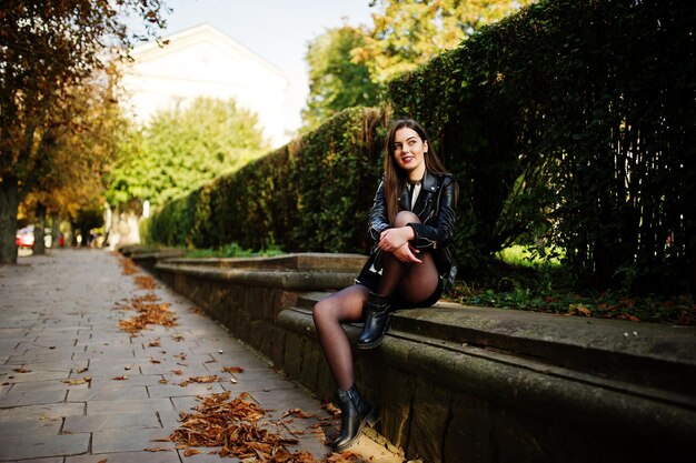 Stylish brunette girl wear on leather jacket and shorts sitting at street of autumn city