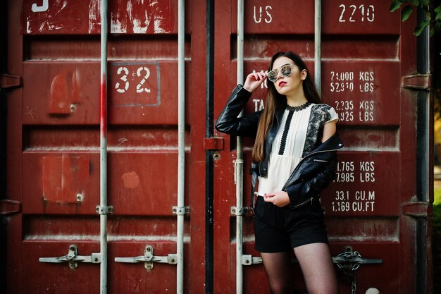Free photo stylish brunette girl wear on leather jacket and shorts sunglasses against shipping container