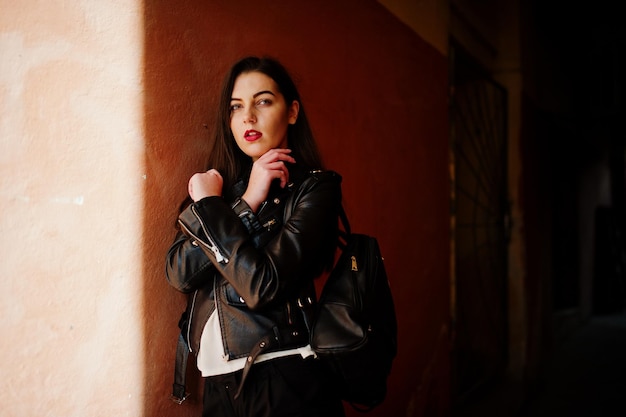 Stylish brunette girl wear on leather jacket and shorts with backpack against orange wall at shadows