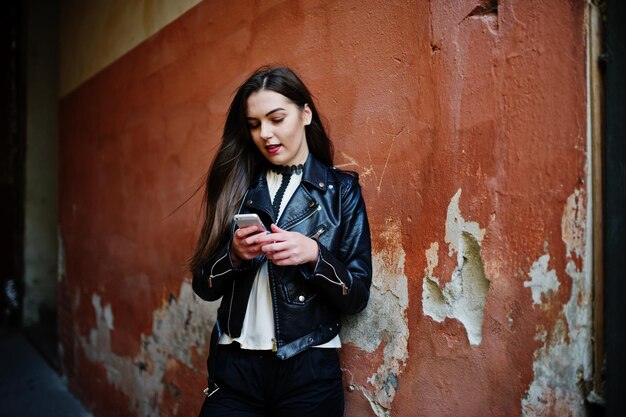 세련된 브루네트 소녀는 가죽 재킷과 반바지를 입고 주황색 벽에 손을 대고 휴대폰을 들고 있다