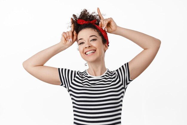 Stylish brunette girl with combed hair and red headband show bull horns gesture and smiling looking stubborn standing against white background