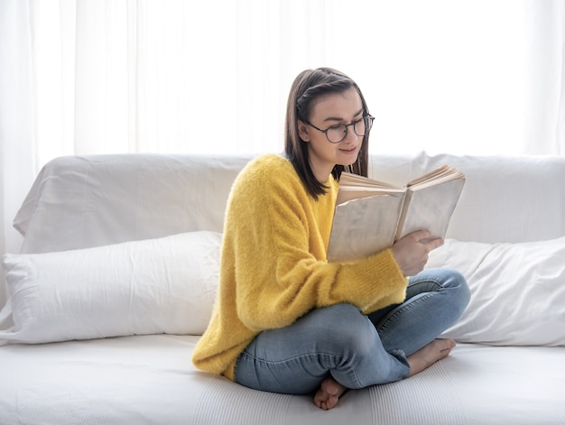Стильная брюнетка в желтом свитере и очках читает книгу дома на диване.