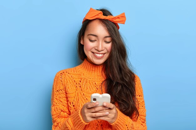 Stylish brunette millennial girl busy checking her email box, holds mobile phone, wears orange headband tied in bow, warm sweater
