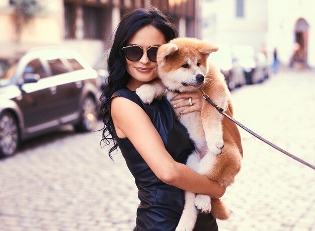 A stylish brunette woman in a black dress and sunglasses, standing on a street and holds a cute purebred puppy Akito.