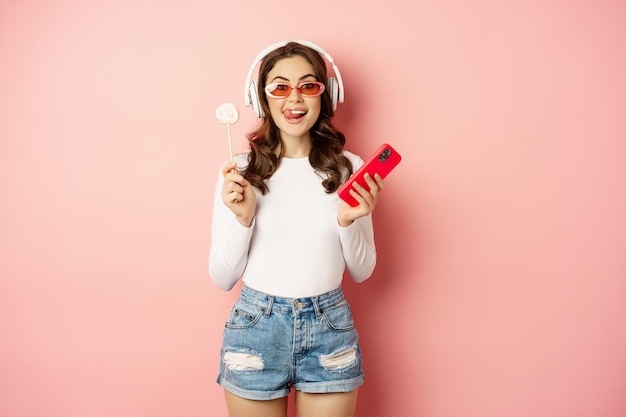 Stylish brunette woman dancing in headphones with mobile phone listening music in earphones licking ...