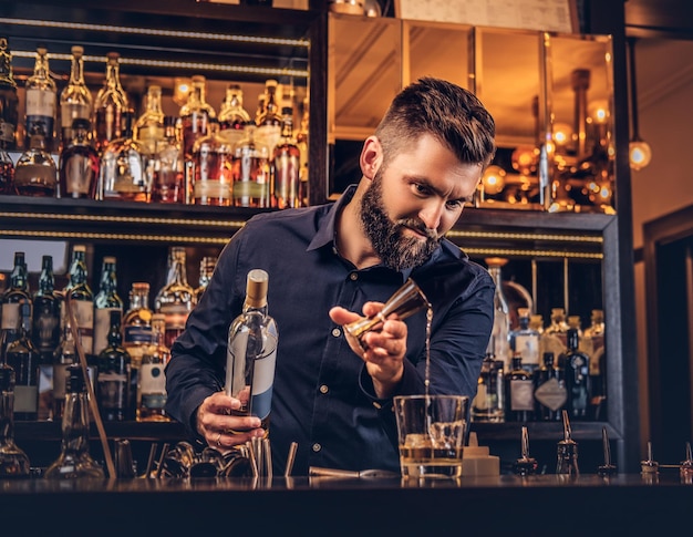 Stylish brutal bartender in a black shirt makes a cocktail at bar counter background.