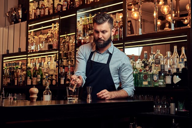 Стильный брутальный бармен в рубашке и фартуке делает коктейль на фоне барной стойки.