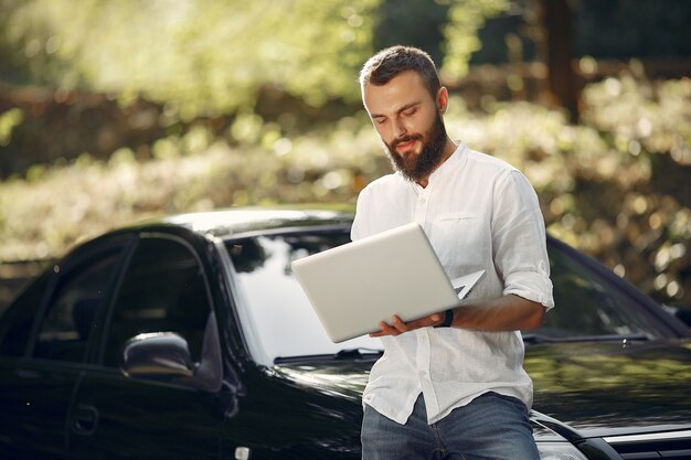 Стильный бизнесмен стоит возле машины и пользуется ноутбуком