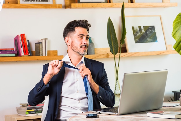 Free photo stylish businessman with laptop and mobile phone on table losing his tie