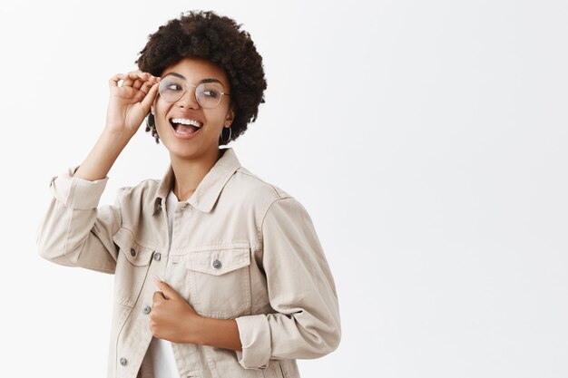 Stylish carefree and confident female dark-skinned woman in glasses and beige shirt, touching clothes and laughing joyfully, looking right, having fun in friends circle