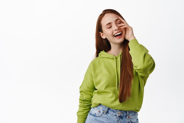 Stylish and carefree redhead natural girl, laughing and smiling carefree, touching glowing clean skin, standing happy on white