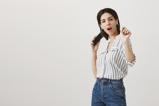 Stylish carefree woman listening music in wireless earphones, dancing with smartphone in hand
