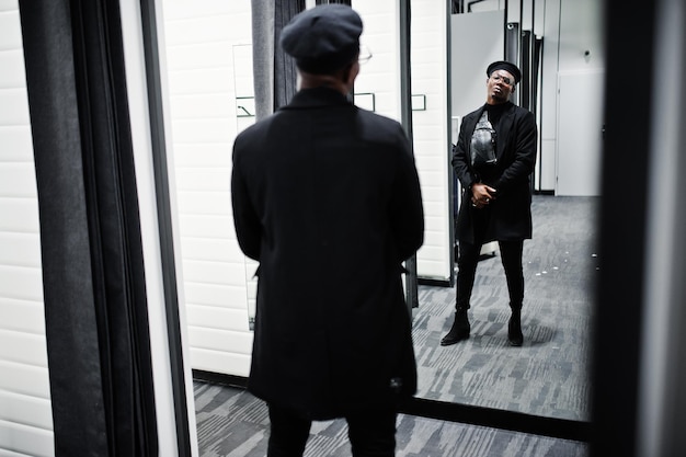 Stylish casual african american man at black beret and overcoat with waist bag at fitting room clothes store looking on mirror