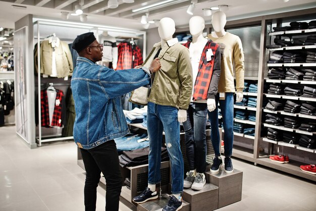 Стильный повседневный афроамериканец в джинсовой куртке и черном берете в магазине одежды смотрит на новую куртку на манекене