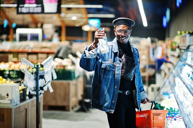 Stylish casual african american man at jeans jacket and black beret holding basket and milk at hand standing near fridge and shopping at supermarket