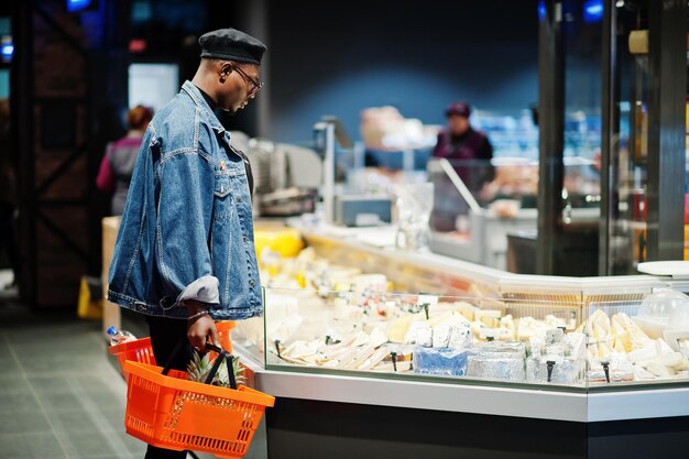 Stylish casual african american man at jeans jacket and black beret holding basket standing near cheese fridge and shopping at supermarket