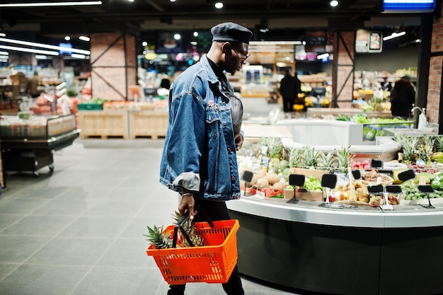 Foto gratuita elegante uomo afroamericano casual in giacca di jeans e berretto nero con cesto con ananas nella sezione frutta biologica del supermercato