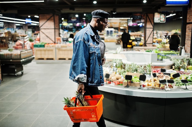 Stylish casual african american man at jeans jacket and black beret holding basket with pineapples in fruits organic section of supermarket