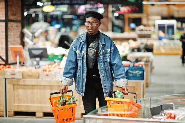 Stylish casual african american man at jeans jacket and black beret holding two baskets walking and shopping at supermarket