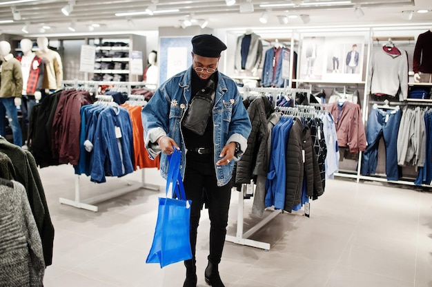ジーンズのジャケットと黒のベレー帽でスタイリッシュなカジュアルなアフリカ系アメリカ人の男、洋服店で青い買い物袋を保持しているファニーパックまたはウエストバッグ