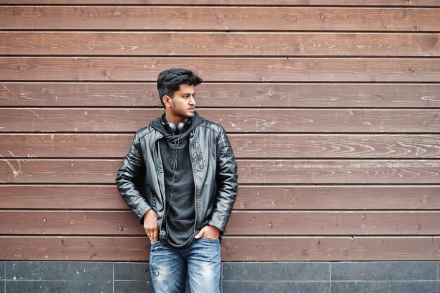 Stylish and casual asian man in black leather jacket headphones posed against wooden wall