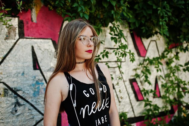 Foto gratuita elegante ragazza hipster casual con occhiali e jeans con cappuccio indossa musica d'ascolto dalle cuffie del telefono cellulare contro il grande muro di graffiti