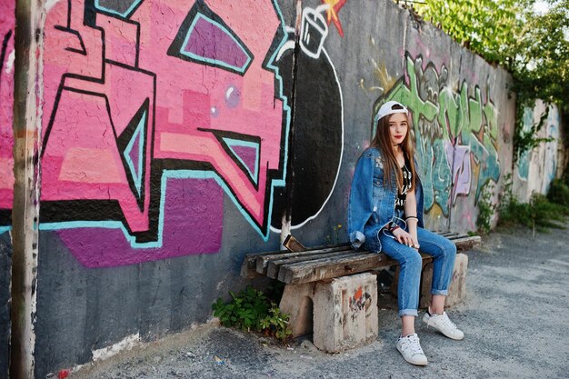Stylish casual hipster girl in cap and jeans wear listening music from headphones of mobile phone against large graffiti wall with bomb