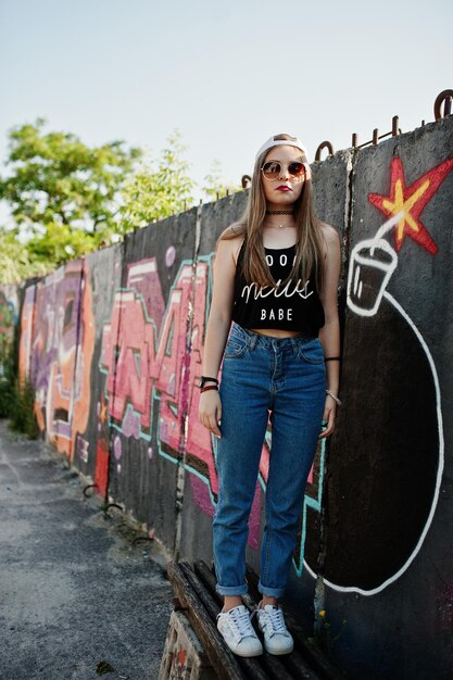 모자 선글라스와 청바지를 입은 세련된 캐주얼 힙스터 소녀는 큰 tnt 폭탄이 있는 큰 낙서 벽에 착용합니다.