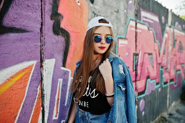 Stylish casual hipster girl in cap sunglasses and jeans wear listening music from headphones of mobile phone against large graffiti wall