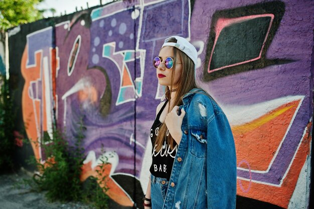 Stylish casual hipster girl in cap sunglasses and jeans wear listening music from headphones of mobile phone against large graffiti wall