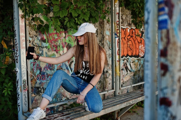 無料写真 キャップとジーンズのスタイリッシュなカジュアルなヒップスターの女の子は、大きな落書きの壁に携帯電話のヘッドフォンから音楽を聴いて着用します