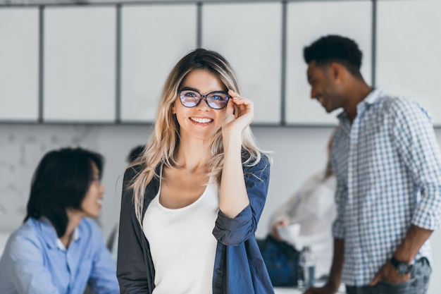 Stylish caucasian female freelancer in black shirt posing in new office while her colleagues talking. Indoor portrait of excited student in glasses having fun after difficult exams with friends.