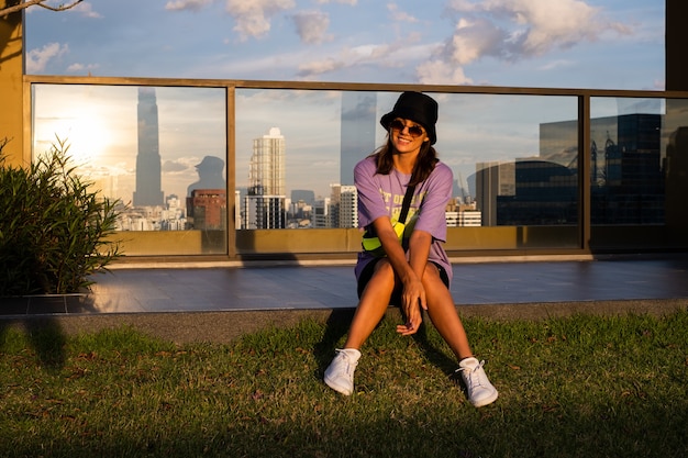 Stylish caucasian woman in trendy panama and waist neon bag on roof in Bangkok.