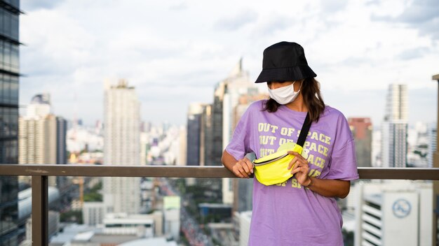 Stylish caucasian woman in trendy panama and waist neon bag on roof in Bangkok