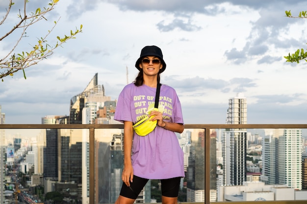 Free photo stylish caucasian woman in trendy panama and waist neon bag on roof in bangkok